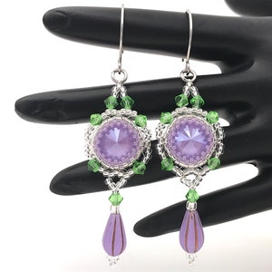 May include: A pair of dangle earrings with a silver tone finish. The earrings feature a large, round, lavender crystal surrounded by smaller, clear crystals and green beads. The earrings are finished with a teardrop-shaped lavender crystal.