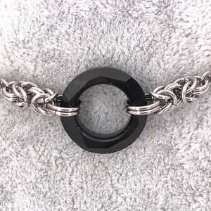Peut inclure: Un collier argenté avec un pendentif noir circulaire et facetté. Le pendentif est relié à une chaîne tissée de chaque côté. Le collier est présenté sur une surface texturée gris clair.
