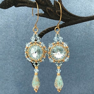 May include: A pair of gold dangle earrings with a teardrop-shaped blue and gold bead at the bottom. The earrings feature a round crystal surrounded by gold and silver beads.
