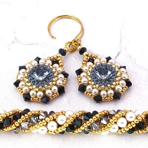 May include: A set of beaded earrings and a bracelet with a gold, black, white, and gray color scheme. The earrings feature a large, faceted, gray crystal surrounded by white pearls and black beads. The bracelet is a braided design with white pearls, black beads, and gold beads.