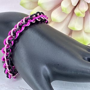 May include: A chainmaille bracelet constructed from interlocking metal rings in alternating colors of vibrant hot pink and glossy black. The bracelet features a repeating pattern of linked circular segments that form a continuous band.