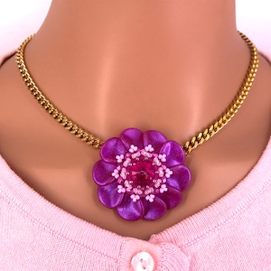 May include: A gold chain necklace with a large purple flower pendant. The flower has a pink center with a clear rhinestone.
