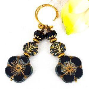 May include: A pair of handcrafted earrings featuring black flower-shaped beads with gold accents. The earrings also include leaf-shaped beads and faceted black beads, all suspended from gold-colored ear wires. The design has a vintage aesthetic.