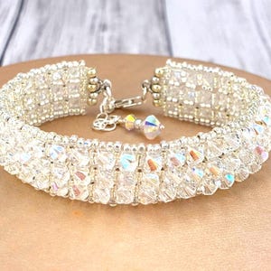 May include: A wide bracelet constructed from multiple rows of clear, faceted crystal beads with an iridescent finish. The piece features a silver-toned metal clasp mechanism and a small dangling crystal charm attached to the closure hardware.