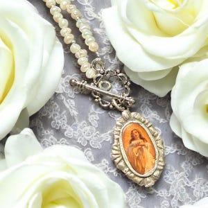 May include: A silver necklace with a white pearl chain and a silver pendant featuring a religious image of a woman in a blue dress. The pendant is surrounded by a silver frame with an ornate design. The necklace is displayed on a white lace background.