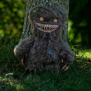 Puede incluir: Una criatura espeluznante, de color marrón oscuro y peluda, con una amplia sonrisa dentuda y ojos anaranjados, posando contra un tronco de árbol. La criatura tiene garras afiladas y está sentada en hierba verde. Una decoración única de Halloween.