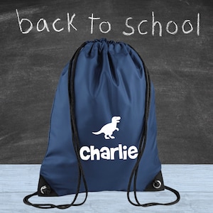 May include: A navy blue drawstring bag featuring a white dinosaur illustration and the name "Charlie". The words "back to school" are written in white chalk on a dark chalkboard. Black drawstring cords.