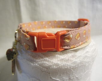 May include: A cat collar with a light pink background and a pattern of candy corn. The collar has an orange plastic buckle and a small gold-colored heart charm that says "Made with love".