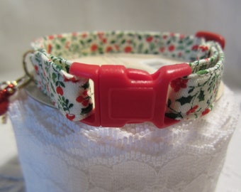 May include: A cat collar with a red plastic buckle and a small gold bell. The collar is patterned with a cream background and a repeating design of red berries and green leaves. The collar is approximately 0.5 inches wide.