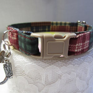 May include: A red, green, and brown plaid cat collar with a beige plastic buckle and a silver charm shaped like a cowboy boot. The charm has the word "Howdy" engraved on it.