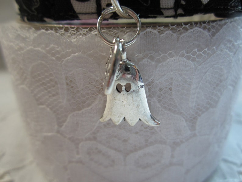 Puede incluir: Un amuleto de plata con forma de fantasma con dos peque&ntilde;os agujeros para los ojos. El amuleto cuelga de una cadena de plata y est&aacute; unido a un fondo de encaje blanco.