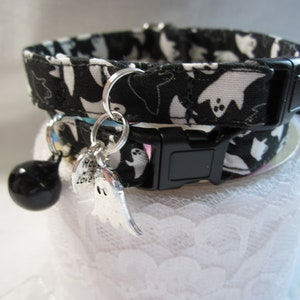 Puede incluir: Collar de gato blanco y negro con un patr&oacute;n de fantasmas. El collar tiene una hebilla negra y un charm de campana plateado con un charm de fantasma adjunto.