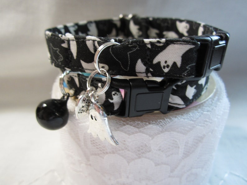Puede incluir: Collar de gato blanco y negro con un patr&oacute;n de fantasmas. El collar tiene una hebilla negra y un charm de campana plateado con un charm de fantasma adjunto.