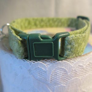 May include: A green patterned cat collar with a dark green plastic buckle and a small silver bell. The collar is made of fabric and is designed to be adjustable. The collar is approximately 0.5 inches wide.