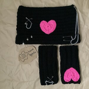 May include: Black crocheted headband, wrist warmers, and a pink heart applique. The headband has a silver chain and safety pins. The wrist warmers also have safety pins. The tag says "hand byleila".