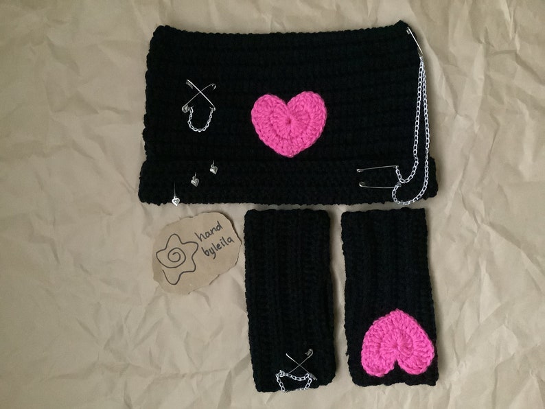 May include: Black crocheted headband, wrist warmers, and a pink heart applique. The headband has a silver chain and safety pins. The wrist warmers also have safety pins. The tag says "hand byleila".