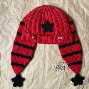 May include: Red and black crocheted hat with ear flaps and star embellishments. The hat has a black star on the top and black stars on the ends of the ear flaps. The hat is handmade by Leila.