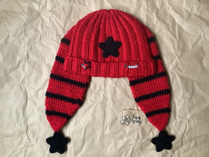 May include: Red and black crocheted hat with ear flaps and star embellishments. The hat has a black star on the top and black stars on the ends of the ear flaps. The hat is handmade by Leila.