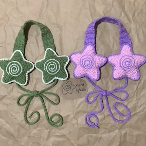 Puede incluir: Dos diademas de ganchillo con orejeras en forma de estrella. Una diadema es verde con remolinos blancos y la otra es morada con remolinos rosas. Ambas diademas tienen un lazo en la parte posterior. La etiqueta dice "hand by leila".