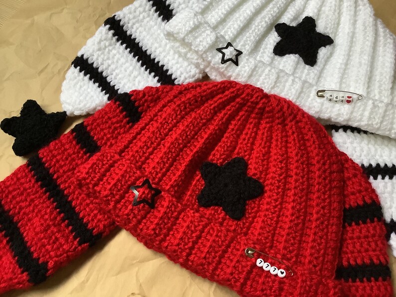 May include: Two crocheted hats with black and white stripes. One hat is red with a black star and the other is white with a black star. Both hats have a safety pin with a number on it.