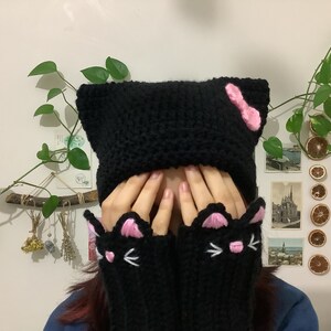 May include: Black knitted cat hat with pink ears and a pink bow. The hat is worn by a person with long brown hair. The person is also wearing black knitted cat mittens with pink ears and white whiskers.