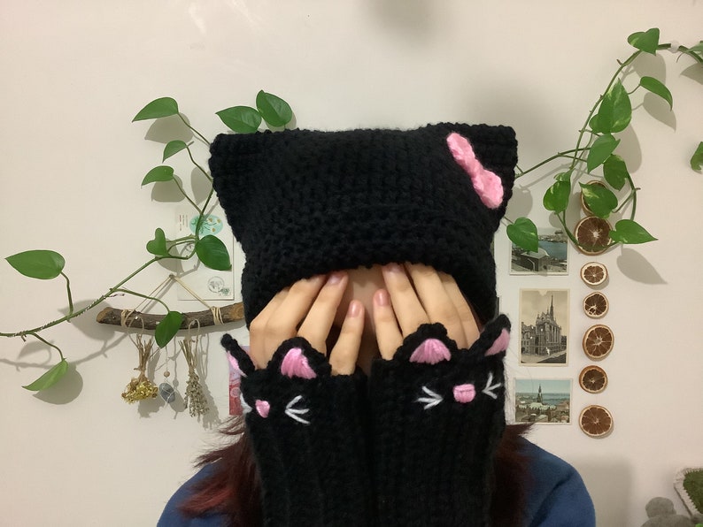 May include: Black knitted cat hat with pink ears and a pink bow. The hat is worn by a person with long brown hair. The person is also wearing black knitted cat mittens with pink ears and white whiskers.