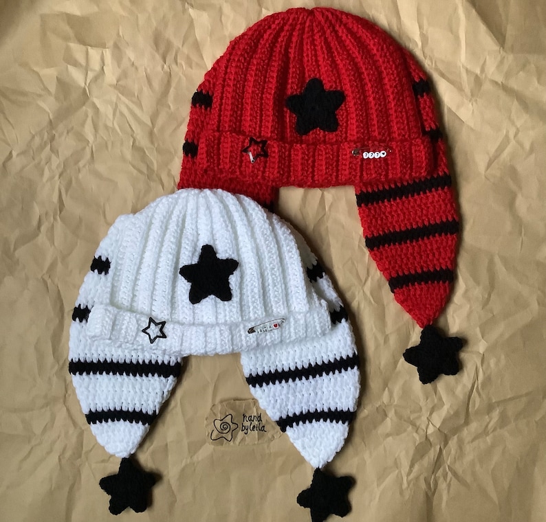 May include: Two crocheted hats with ear flaps. One hat is red with black stripes and a black star. The other hat is white with black stripes and a black star. Both hats have a black star on the top of the ear flap. The hats are on a brown paper background. Hand by Leila.