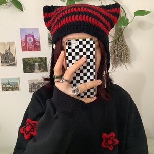 May include: A person wearing a black and red striped knit hat with cat ears. The hat has a black pom-pom on top. The person is also wearing a black sweater with red embroidered flowers. The person is holding a black and white checkered phone case.
