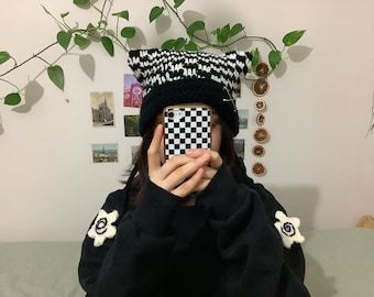 Könnte beinhalten: Eine Person trägt eine schwarz-weiß karierte Strickmütze mit Katzenohren und einen schwarzen Pullover mit weißen gestickten Blumen. Die Person hält eine schwarz-weiß karierte Handyhülle vor ihrem Gesicht.