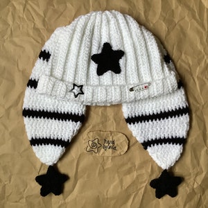 May include: White crocheted hat with black stripes and black star embellishments. The hat has ear flaps with black stars at the ends. The hat has a tag that says "hand by leila".