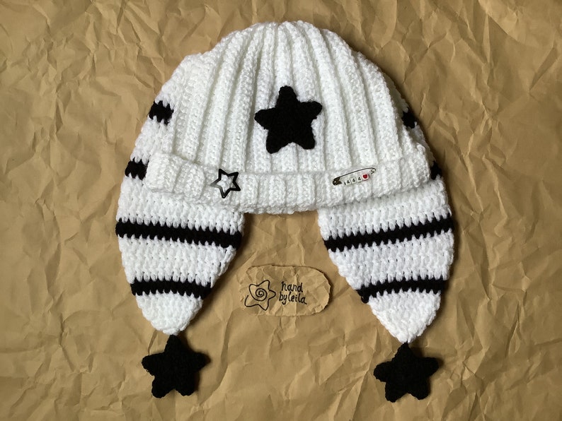 May include: White crocheted hat with black stripes and black star embellishments. The hat has ear flaps with black stars at the ends. The hat has a tag that says "hand by leila".
