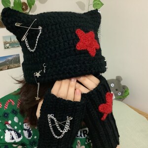 May include: Black crocheted beanie with cat ears and red star accents. The beanie is decorated with safety pins and chains. Matching black fingerless gloves with red heart accents.