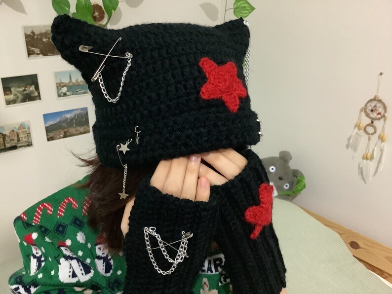 May include: Black crocheted beanie with cat ears and red star accents. The beanie is decorated with safety pins and chains. Matching black fingerless gloves with red heart accents.
