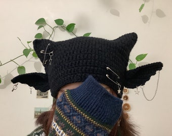 May include: Black crocheted hat with bat wings, safety pins, and star charms. The hat is worn by a person with a blue and green patterned scarf.