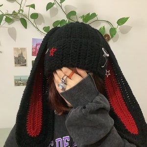 May include: Black crochet beanie with long red and black bunny ears. The beanie has a star design and safety pins attached.