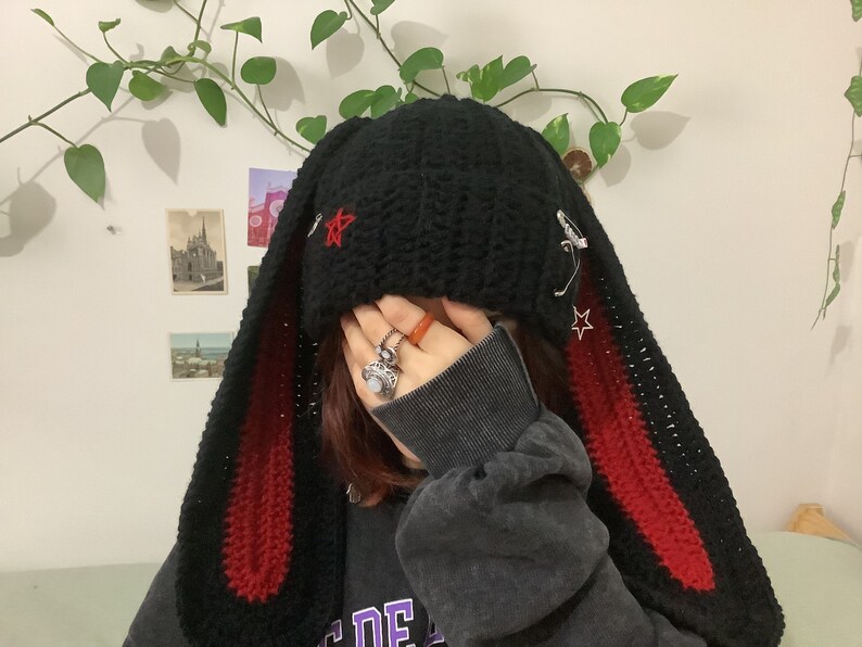 May include: Black crochet beanie with long red and black bunny ears. The beanie has a star design and safety pins attached.