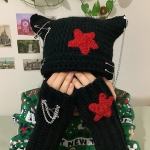 May include: Black crocheted beanie with red star and safety pin accents. The beanie has a chain attached to it. The beanie is being worn by a person wearing a green and red sweater.