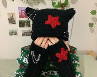 May include: Black crocheted beanie with red star and safety pin accents. The beanie has a chain attached to it. The beanie is being worn by a person wearing a green and red sweater.