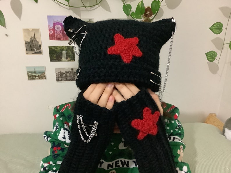 May include: Black crocheted beanie with red star and safety pin accents. The beanie has a chain attached to it. The beanie is being worn by a person wearing a green and red sweater.