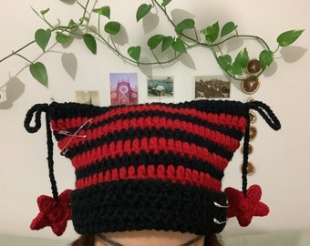 Könnte beinhalten: Eine gestrickte Mütze mit schwarzen und roten Streifen und zwei roten sternförmigen Verzierungen an den Seiten. Die Mütze hat ein schwarzes Band um den unteren Rand und wird mit Sicherheitsnadeln befestigt.