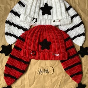 May include: Two crocheted hats with black and white stripes and black star embellishments. The hats have long ear flaps with black pom-poms. One hat is white and the other is red. The hats have a tag that says "hand by leila".
