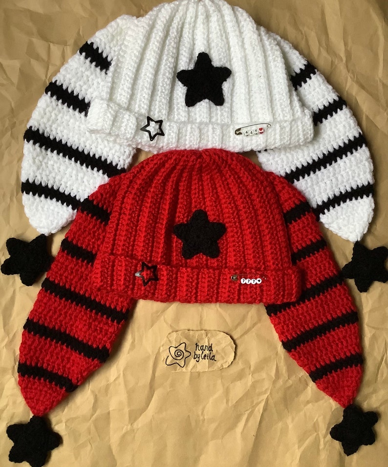 May include: Two crocheted hats with black and white stripes and black star embellishments. The hats have long ear flaps with black pom-poms. One hat is white and the other is red. The hats have a tag that says "hand by leila".