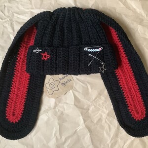 May include: Black beanie with red and black ear flaps. The beanie has a star and a safety pin on it. The text "hand by leila" is on a tag attached to the beanie.
