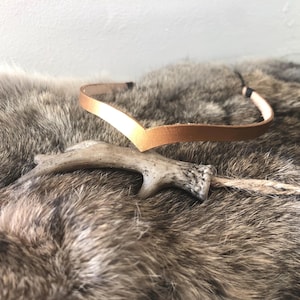 May include: A brown leather headband with a pointed design, accented with a white antler. The headband is lying on a brown fur surface.