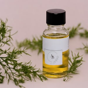 Puede incluir: Una botella de vidrio transparente con una tapa negra llena de un líquido amarillo. La botella tiene una etiqueta blanca con el texto "Where Herbalism Meets Beauty" y un logotipo con las letras "AB" dentro de un círculo. El texto "Est. 2019" está debajo del logotipo.
