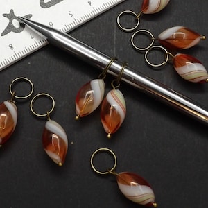 May include: Close-up of several handmade knitting stitch markers. Each marker features a teardrop-shaped, translucent bead with brown, white, and cream stripes, suspended from a gold-coloured ring. A silver knitting needle and a ruler are also in the image.
