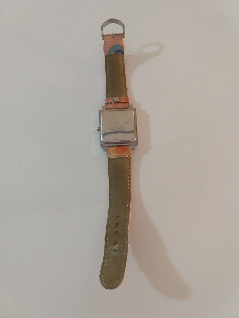 May include: A silver square watch with a brown leather band. The watch face has a white background with black text that reads "Alberto Fiore" and "T.I.M.E.  MADE IN CHINA".
