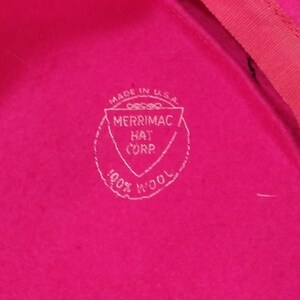 May include: Close-up of a bright pink hat with a red trim. The hat features a white stamp that reads "Made in U.S.A. MERRIMAC HAT CORP. 100% WOOL."