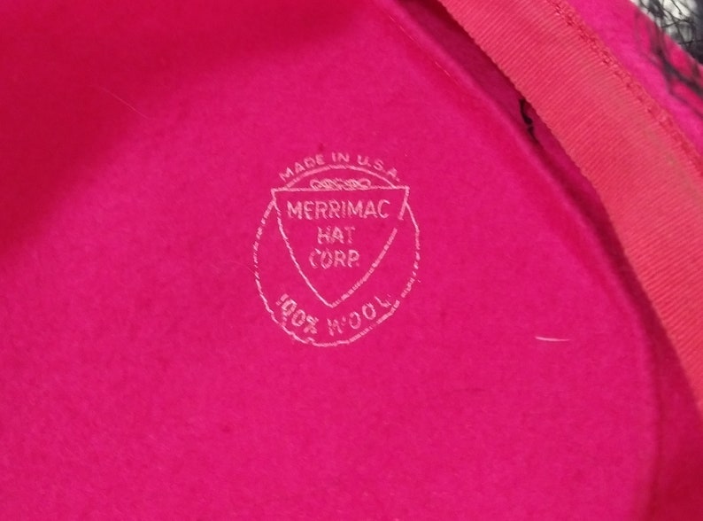 May include: Close-up of a bright pink hat with a red trim. The hat features a white stamp that reads "Made in U.S.A. MERRIMAC HAT CORP. 100% WOOL."