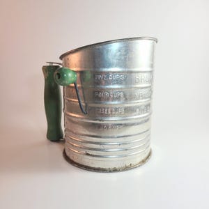 May include: A vintage metal flour sifter with a green wooden handle and a matching green crank knob. The cylindrical body features embossed measurement markings for one, three, four, and five cups, along with the text BROOKS MEASURE GUARANTEED.